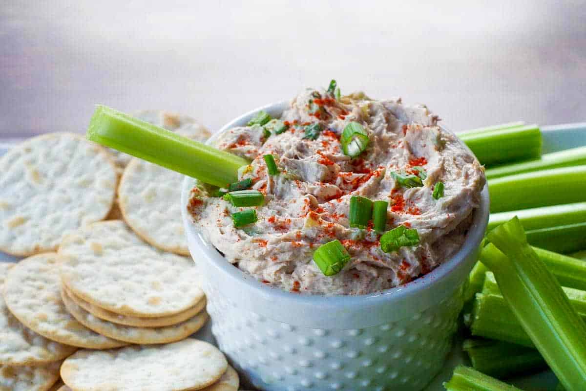 tuna dip in white container on a tray with crackers and celery with a piece of celery sitting in the dip