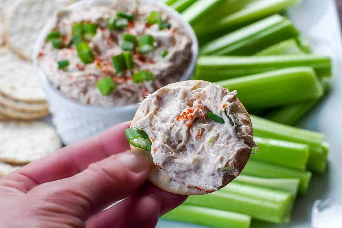 tuna dip in white container on a tray with crackers and celery with a hand holding up a cracker with tuna dip on it