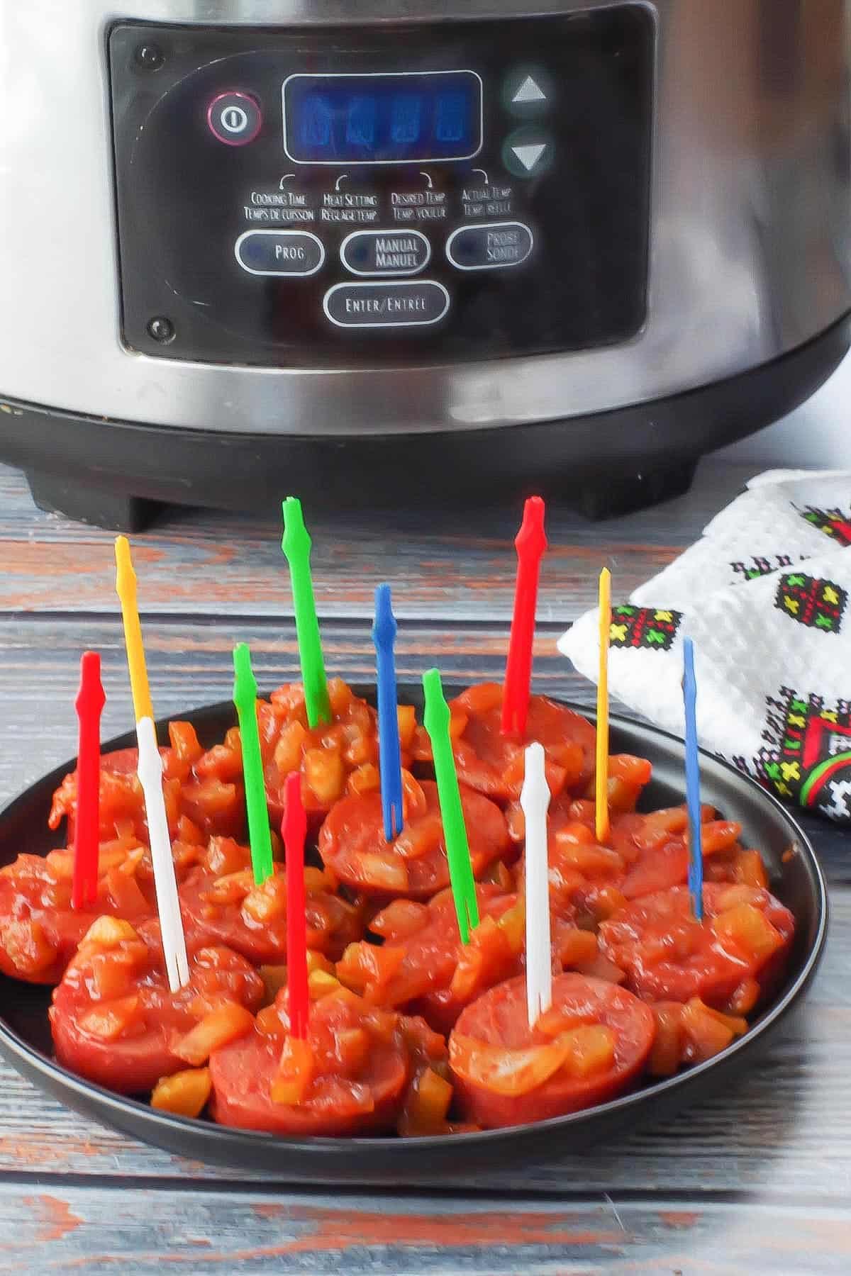 slow cooker kielbasa appetizer with colorful plastic skewers, on a black round plate with a slow cooker in the background
