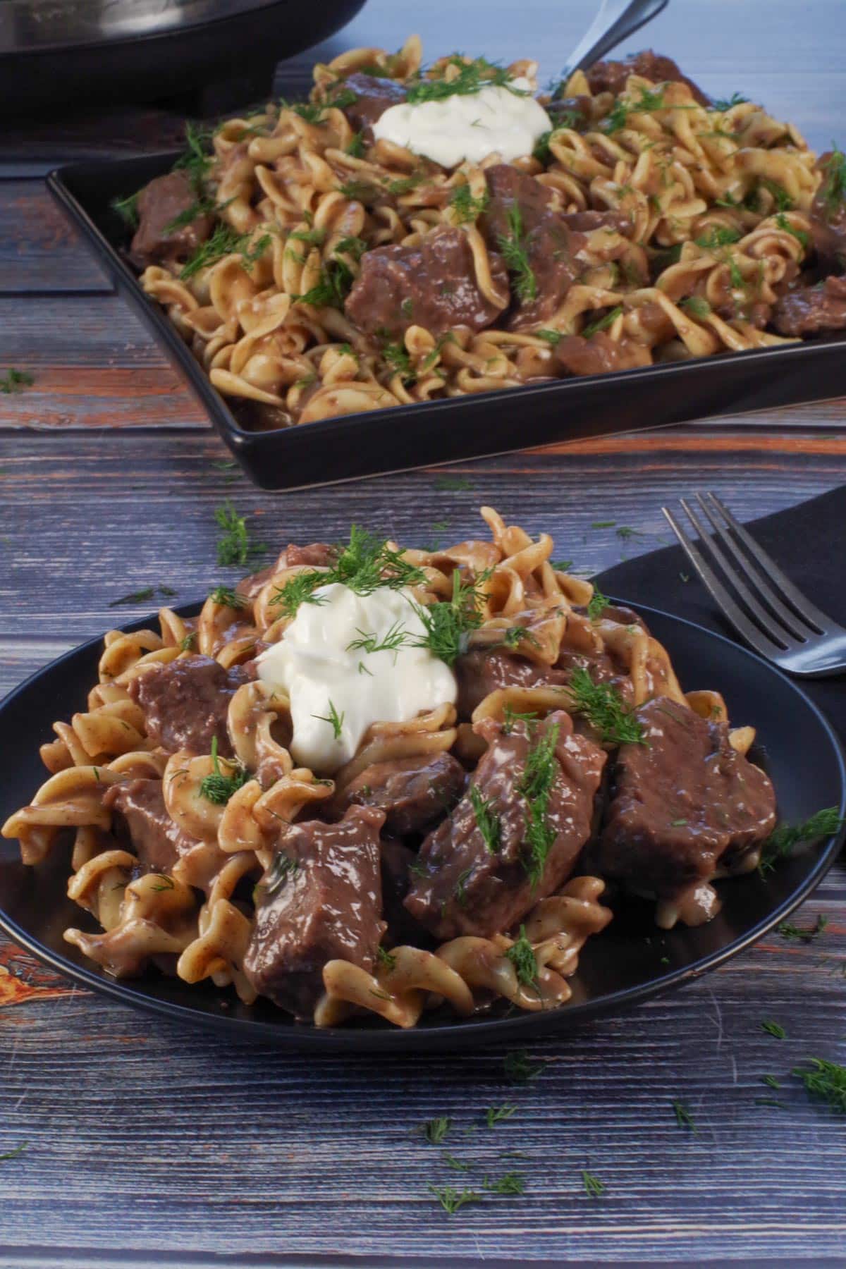 beef stroganoff on a black plate with a tray of more in the background