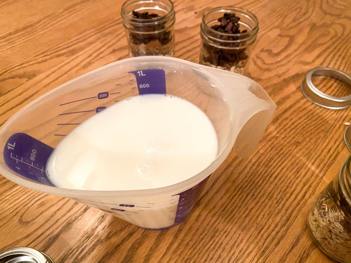 milk in a measuring cup with jars of oat mixture in background, on a table,