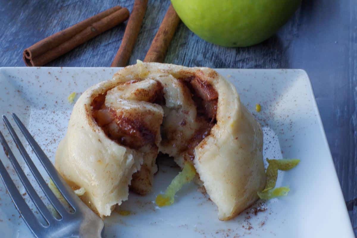 1 cinnamon roll, cut open, on a white plate with lemon zest and cinnamon sticks and a green apple in the background