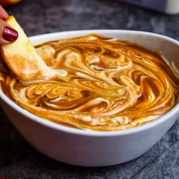 caramel apple cheesecake dip in a white bowl, with a slice of apple being dipped