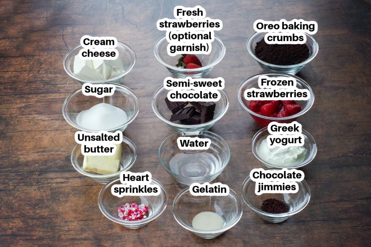 ingredients in Valentine's Day Cheesecake in glass bowls, labelled, on a dark brown faux wood surface
