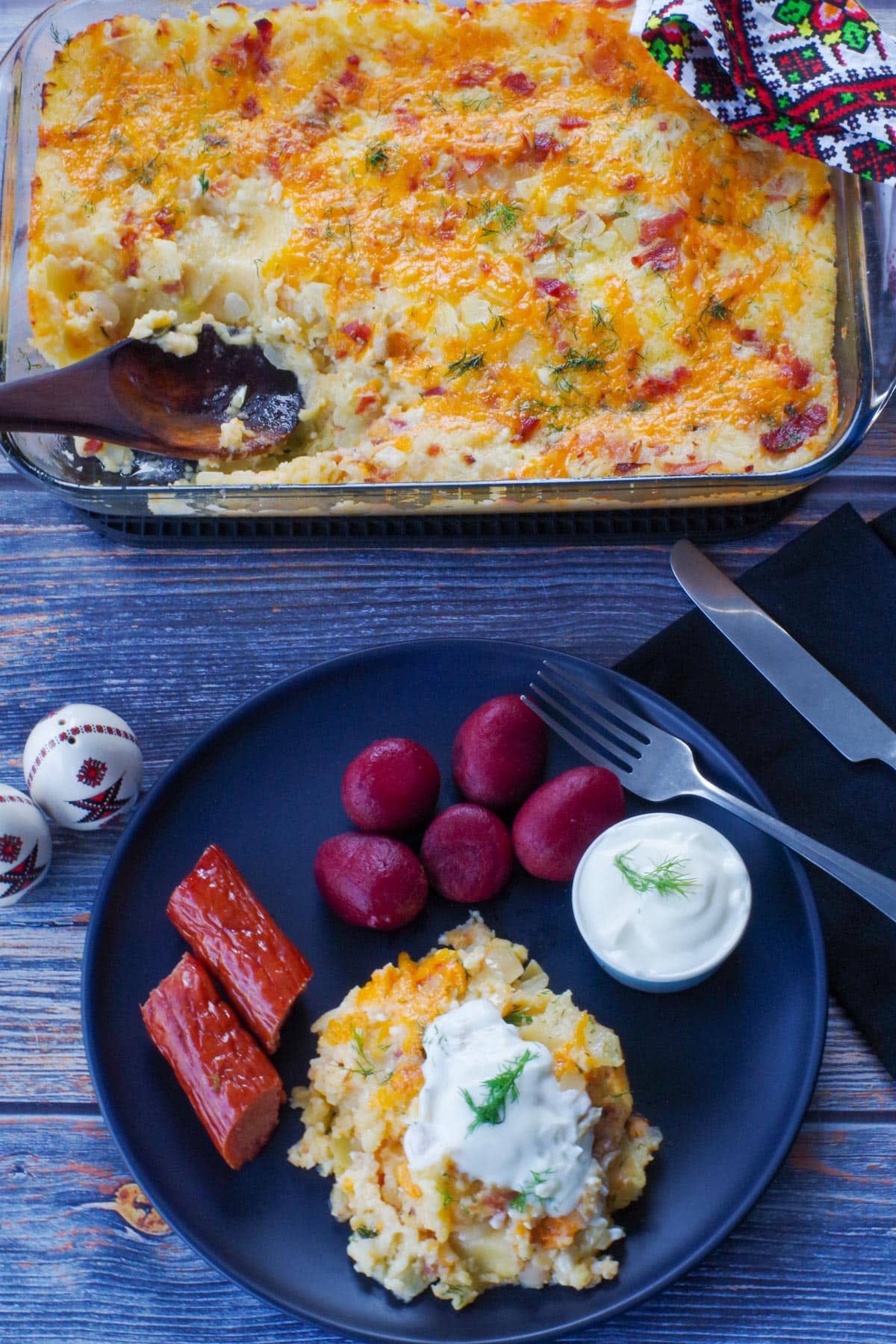 pierogi casserole on a black plate with beets, kielbasa and sour cream, with more casserole in the background
