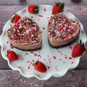 2 Valentine's Day Chocolate Strawberry Cheesecakes on a white cake platter with strawberries and heart sprinkles