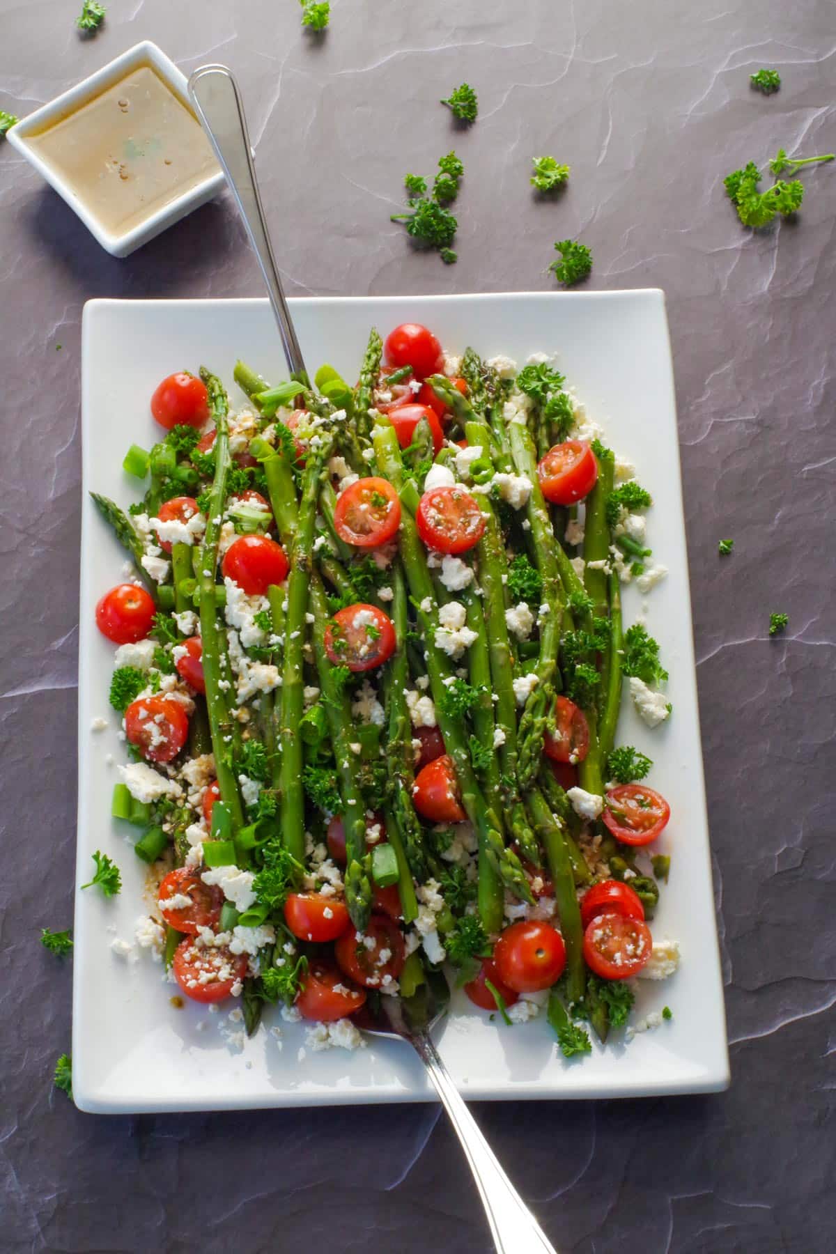 asparagus and tomato salad on a white platter with dressing in the background