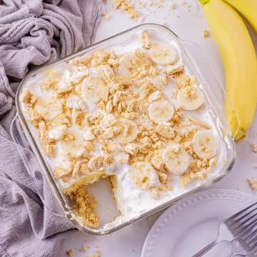 Banana dessert recipe in a glass 8x8 pan, with a piece missing, and bananas on one side and mauve linen on the other side