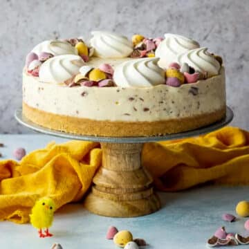 Mini Egg cheesecake on a wooden cake stand with yellow linen behind and Easter decorations (little chicks and eggs) in front