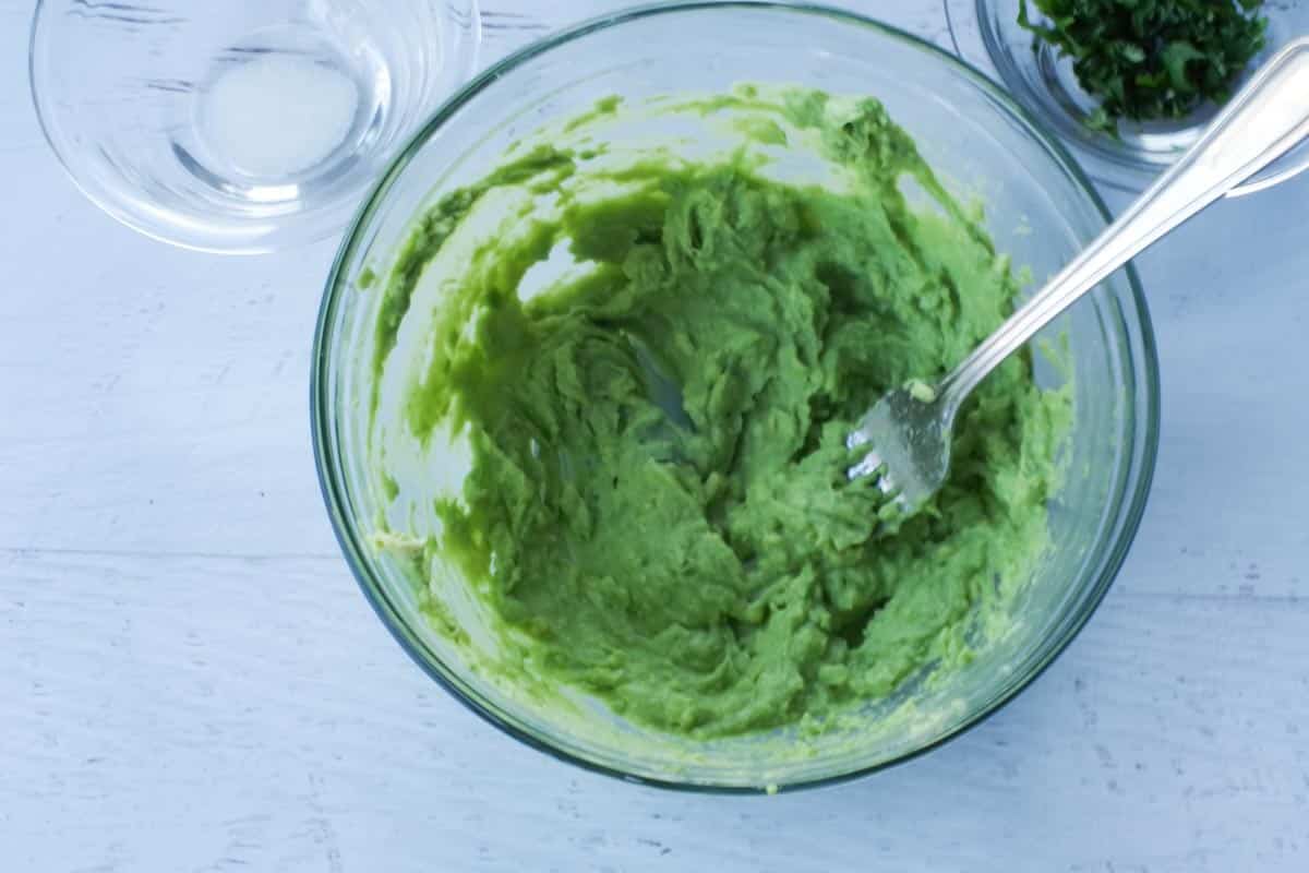 avocado being mashed with a fork, in a glass bowl
