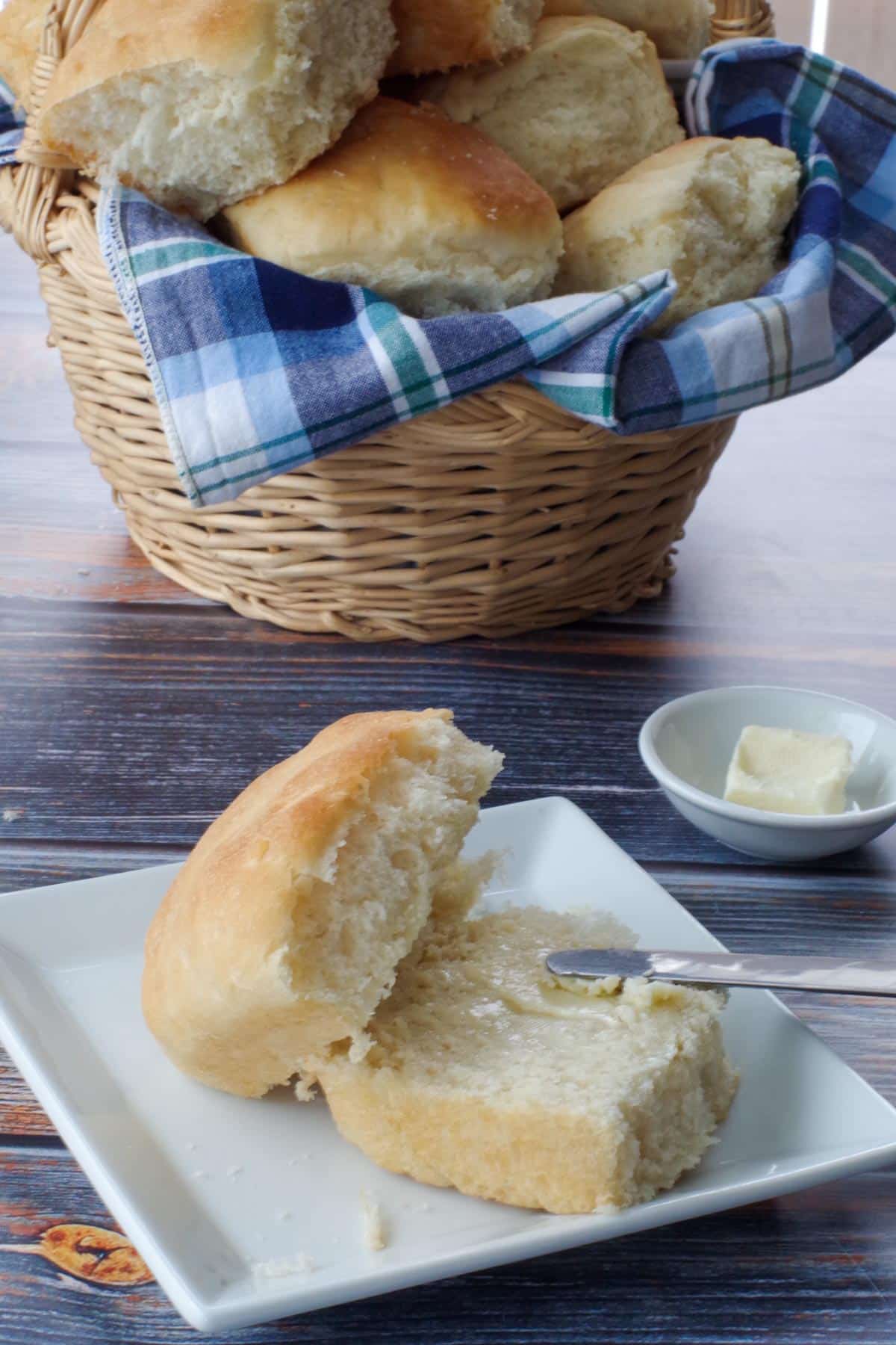 This image shows a roll on a plate with butter. Behind the plate is a basket filled with rolls. 