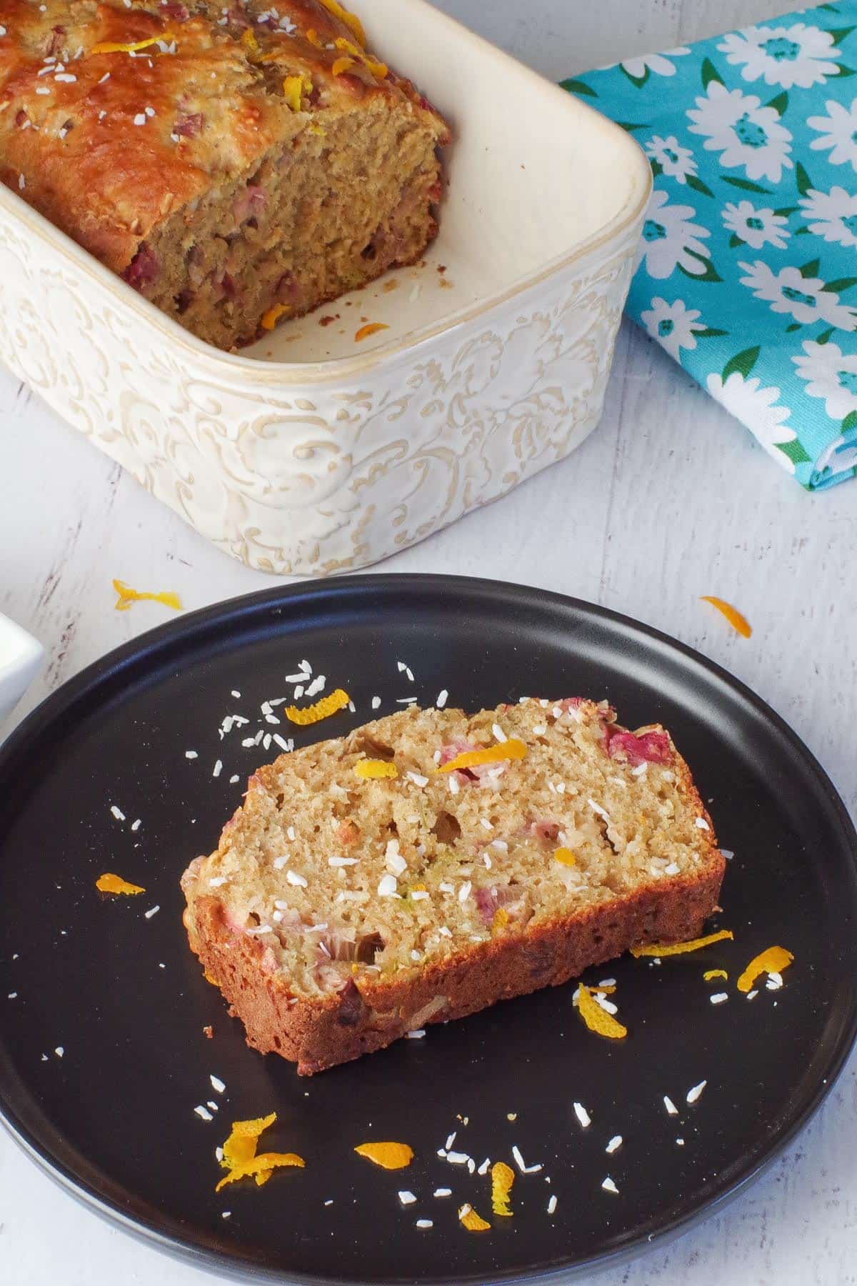 piece of healthy rhubarb bread on a black plate with a ½ a loaf of rhubarb bread in a pan in the back