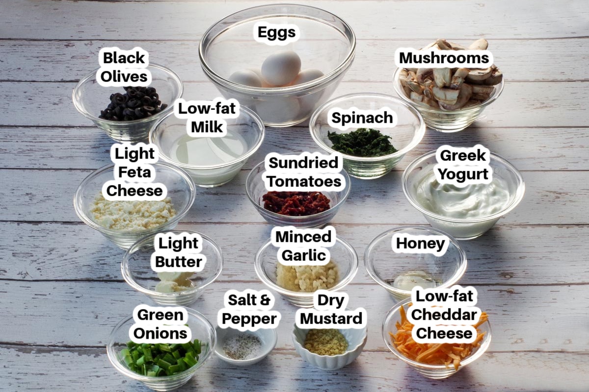 Ingredients needed for Mediterranean ww quiche in glass bowls, labelled on a faux white wood surface