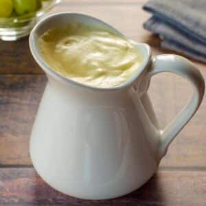 Healthy Hollandaise sauce in a large white creamer, with a bowl of green grapes in background and grey linen