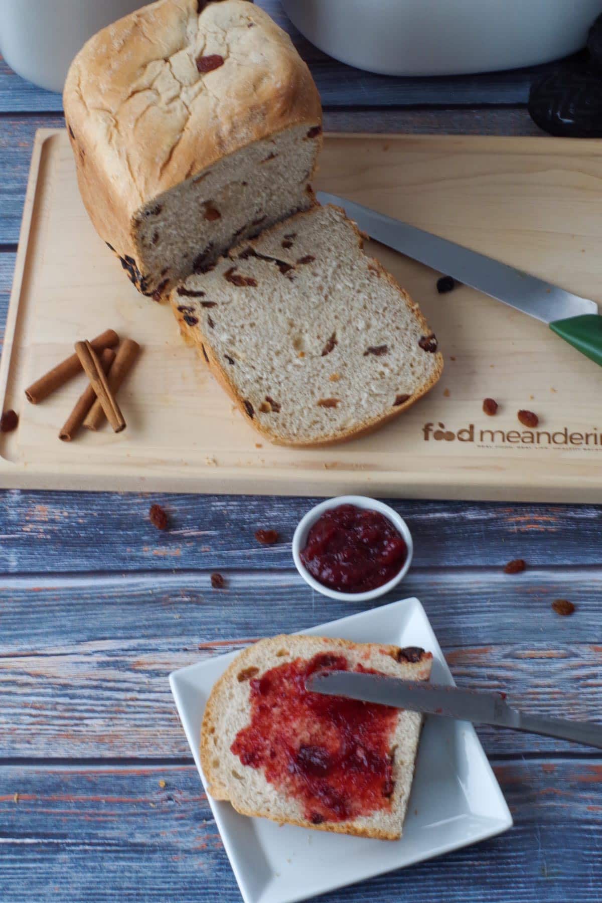 a slice of cinnamon raisin bread machine bread on a white plate in front of a cutting board with a loaf of cinnamon raisin bread, with a piece sliced off