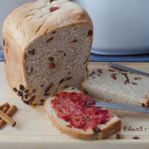 a slice of cinnamon raisin bread machine bread cut in half, with on side spread with jam, in front of a partial loaf of bread, on a cutting board