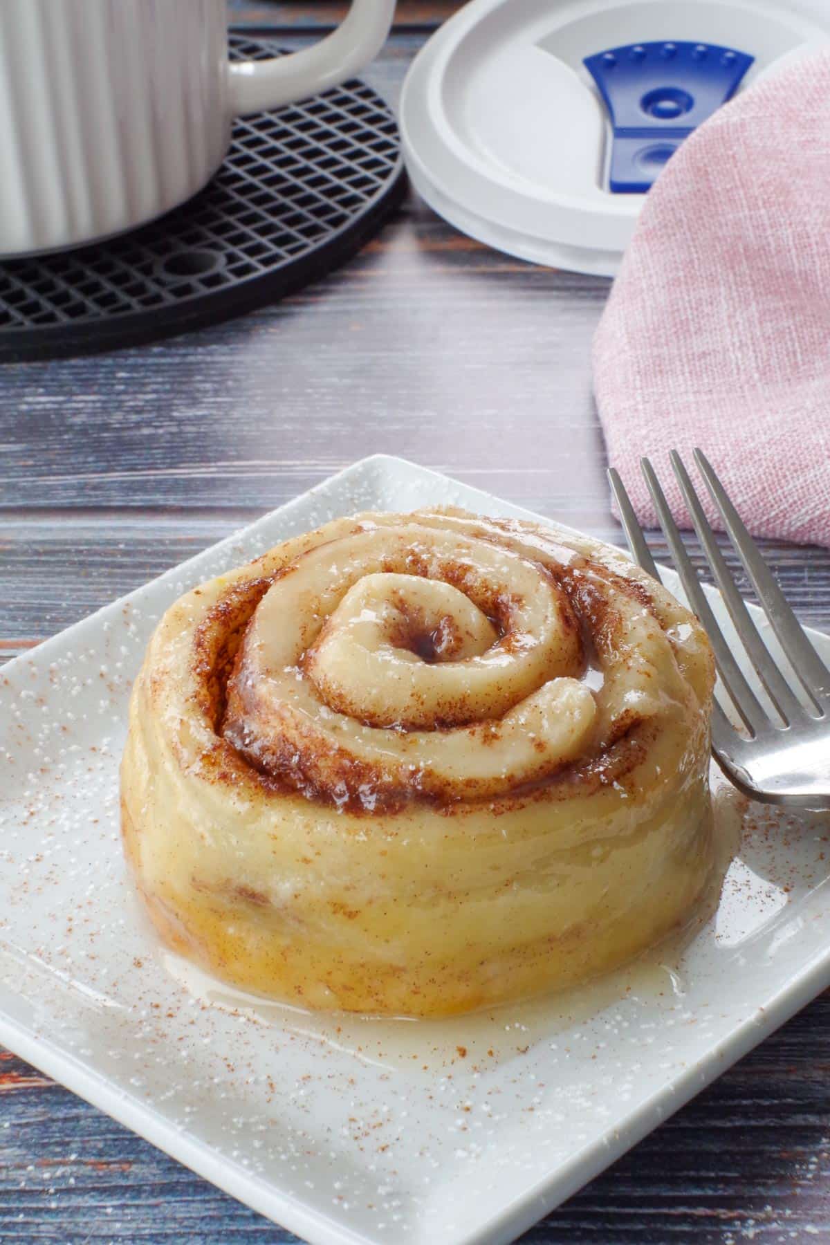 cinnamon roll on a white square plate with a fork, and a meal mug and pink napkin in the background