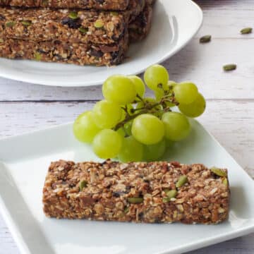 A single granola bar on a white plate with green grapes and a pile of granola bars in the background on a white plate