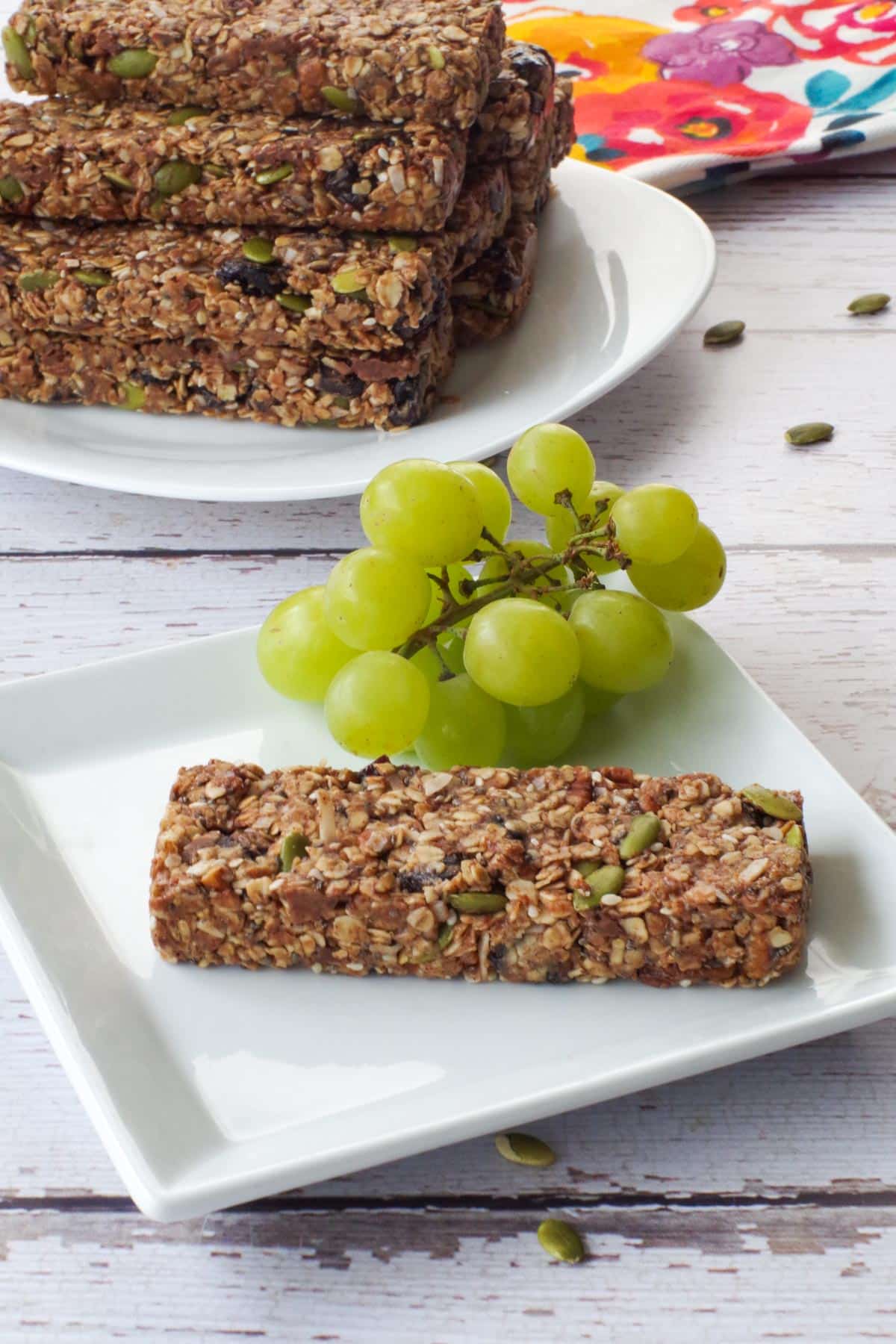 A single granola bar on a white plate with green grapes and a pile of granola bars in the background on a white plate