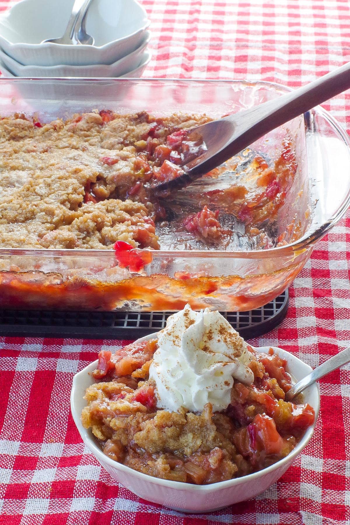 rhubarb crumble topped with whipped cream,  in a white petal dish with a spoon and a glass pan of more rhubarb crisp in the background