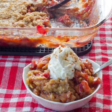 rhubarb crumble topped with whipped cream, in a white petal dish with a spoon and a glass pan of more rhubarb crumble in the background