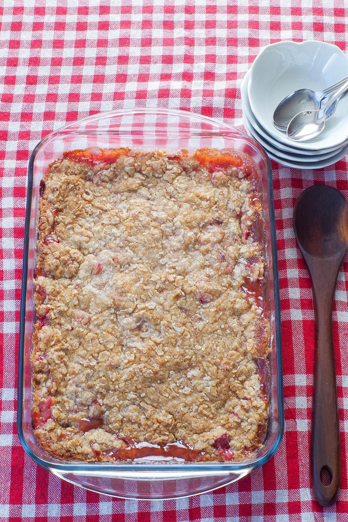 whole rhubarb crisp in glass pan with wooden spoon on one side and white bowls and spoons on the other