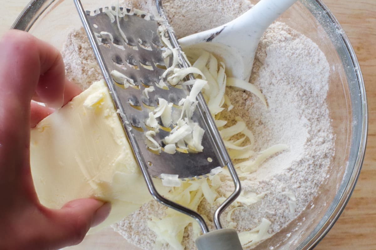 butter being grated into dry ingredients in glass bowl with cheese grater