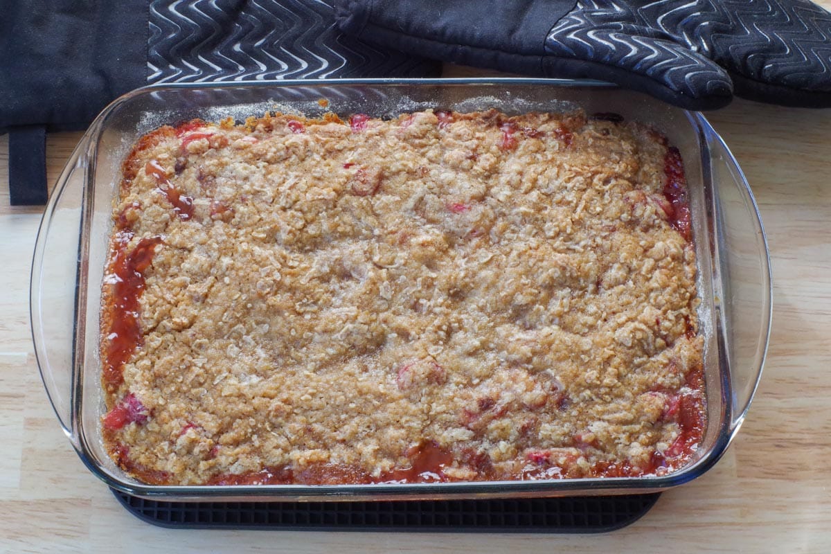 Baked Rhubarb Crumble in a glass pan on a rectangular black trivet, with black oven mitts in the background