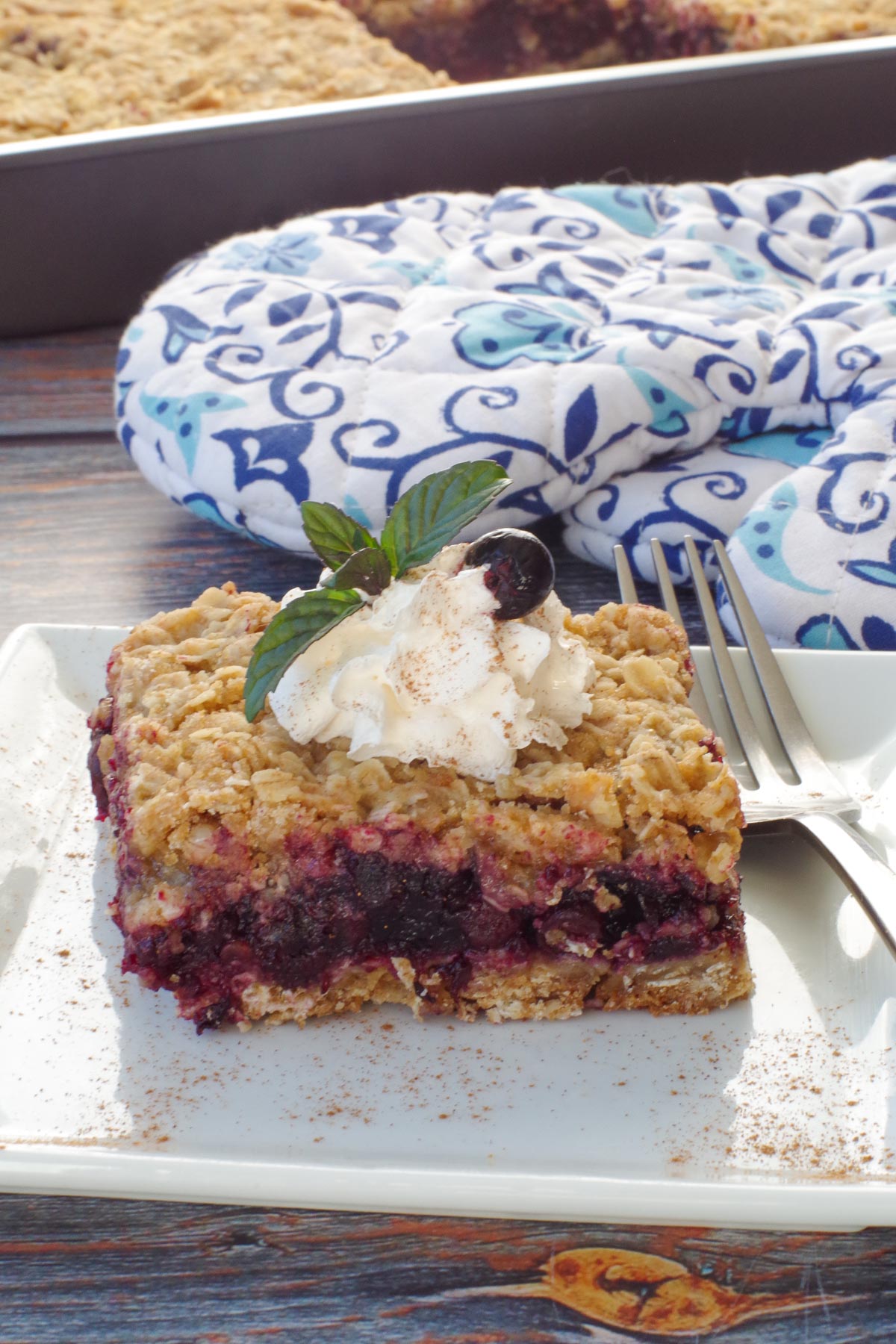 Saskatoon berry square with whipped cream on a white plate with blue flowered oven mitts and a pan of more square in the background