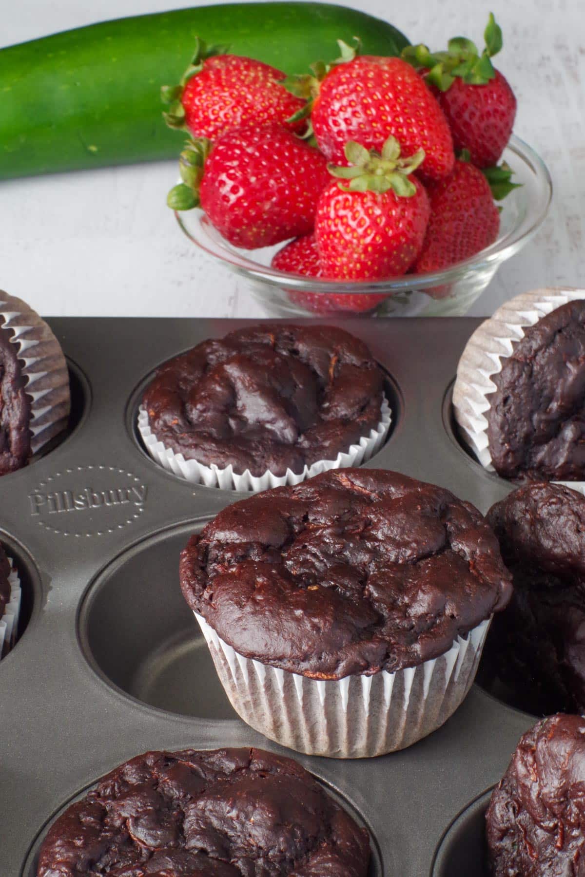 Chocolate Weight Watchers Zucchini Muffins in a muffin tin , with one on top and strawberries and a zucchini in the background