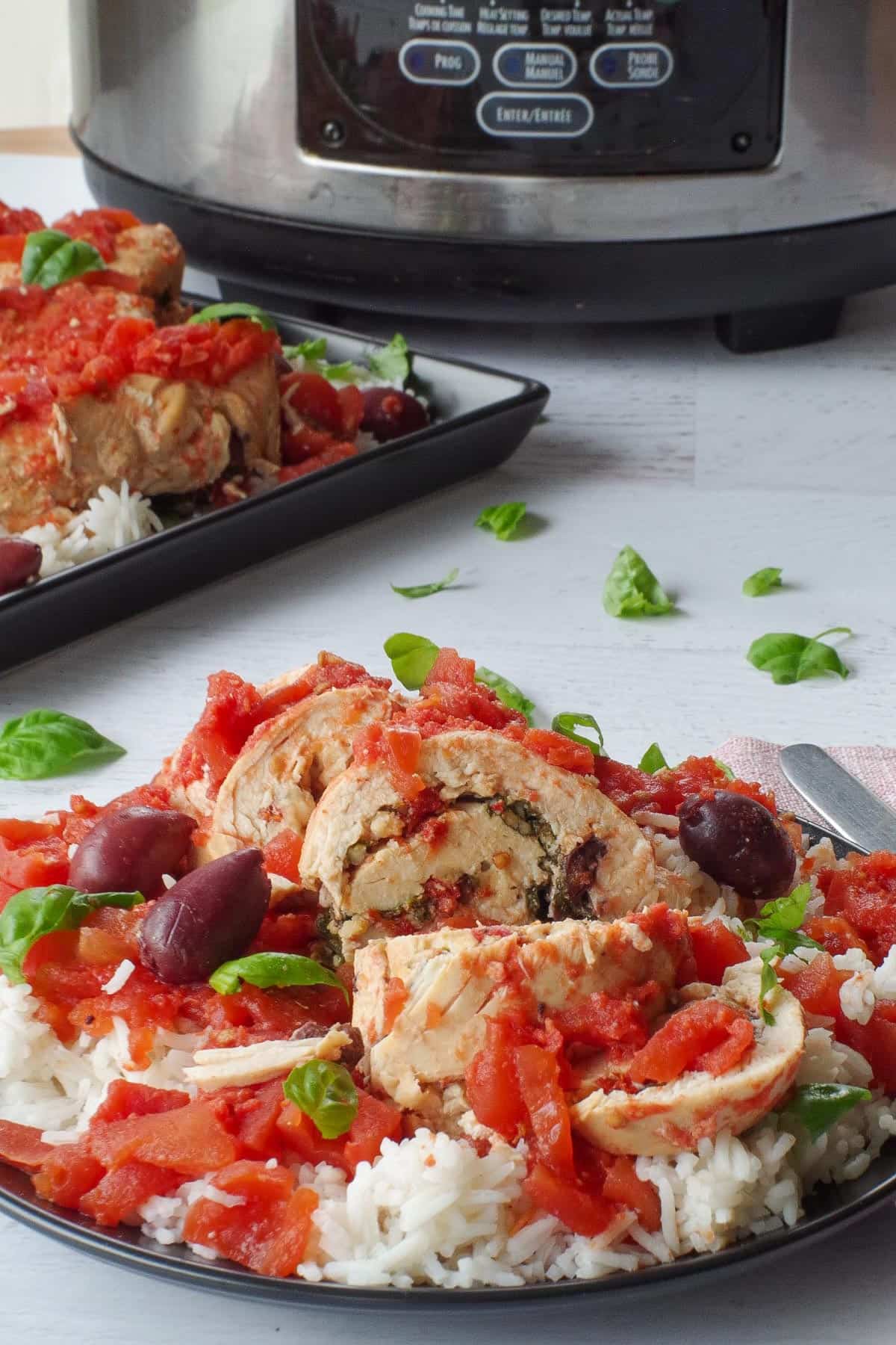 Mediterranean Slow Cooker Stuffed Chicken breast on rice, on a black plate with basil leaves, a platter of more slow cooker Mediterranean chicken breast and a slow cooker, in the background