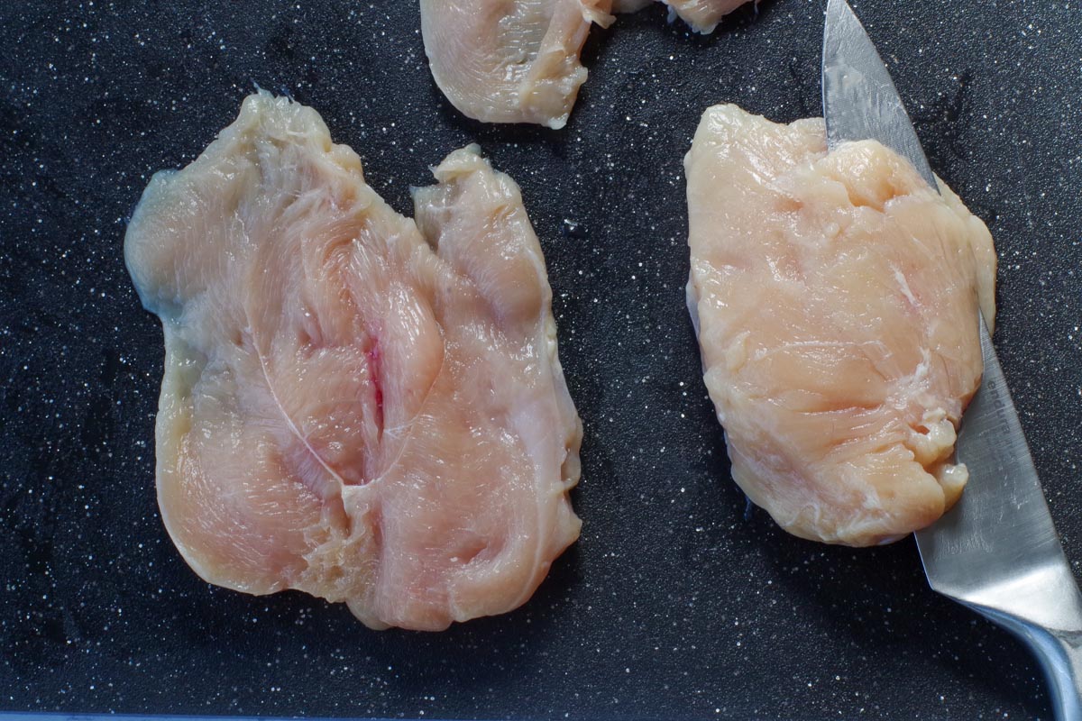 chicken being sliced with a knife on the cutting board, with another piece of raw chicken breast butterflied