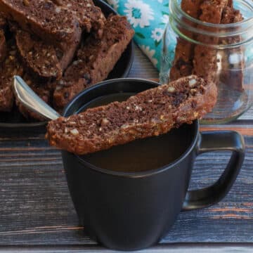 a piece of chocolate zucchini biscotti sitting on a black coffee mug, with a spoon in it, with a plate of more biscotti in the background