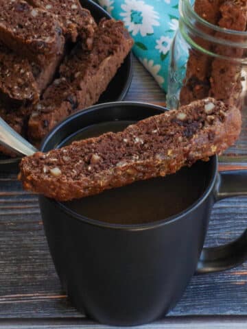 a piece of chocolate zucchini biscotti sitting on a black coffee mug, with a spoon in it, with a plate of more biscotti in the background