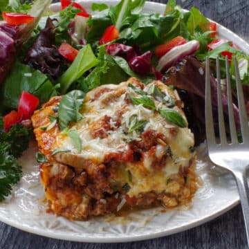 a slice of Zucchini Ground beef lasagna on a white plate with salad and a fork