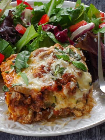 a slice of Zucchini Ground beef lasagna on a white plate with salad and a fork