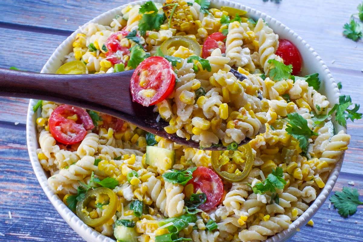 Grilled Mexican Street Corn Pasta Salad in a white bowl with a dark brown wooden spoon holding up some salad