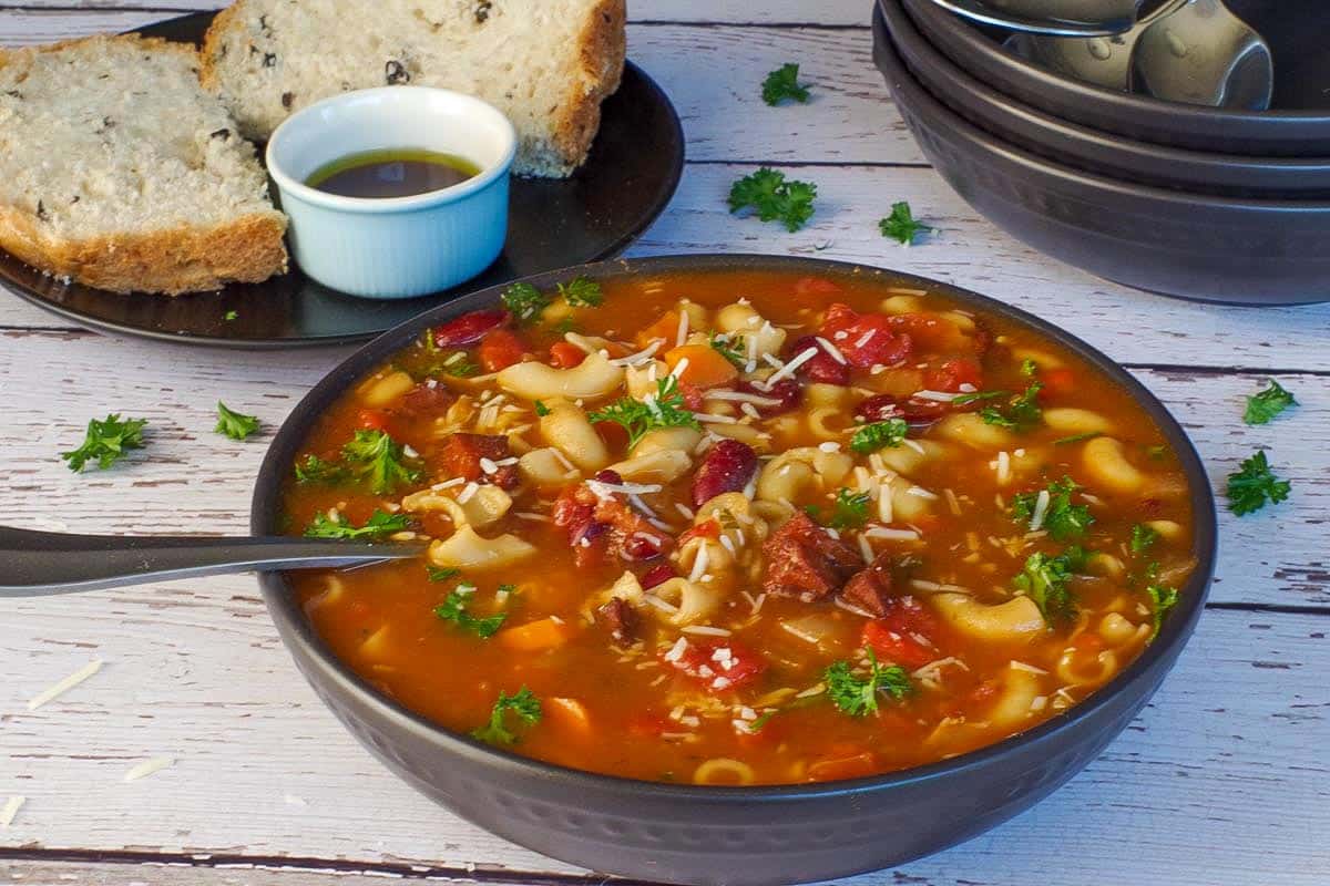 leftover ham macaroni soup in a black bowl, with more black bowls in the background and onion and olive bread with bread and oil