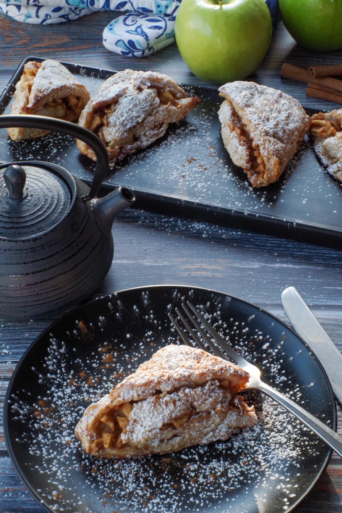 apple cinnamon scone on a black plate with a pot of tea, a tray of more scones and 2 green apples in the background