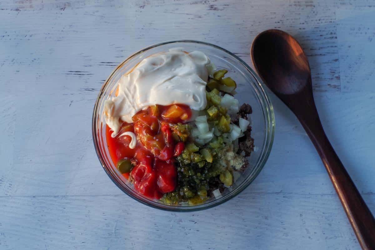 Chopped pickles, onions, and Big Mac dressing (light Catalina salad dressing, mayonnaise, ketchup, relish, onion powder, salt, and vinegar) added to medium glass bowl with cooked ground beef., with a dark brown wooden spoon beside the bowl. 