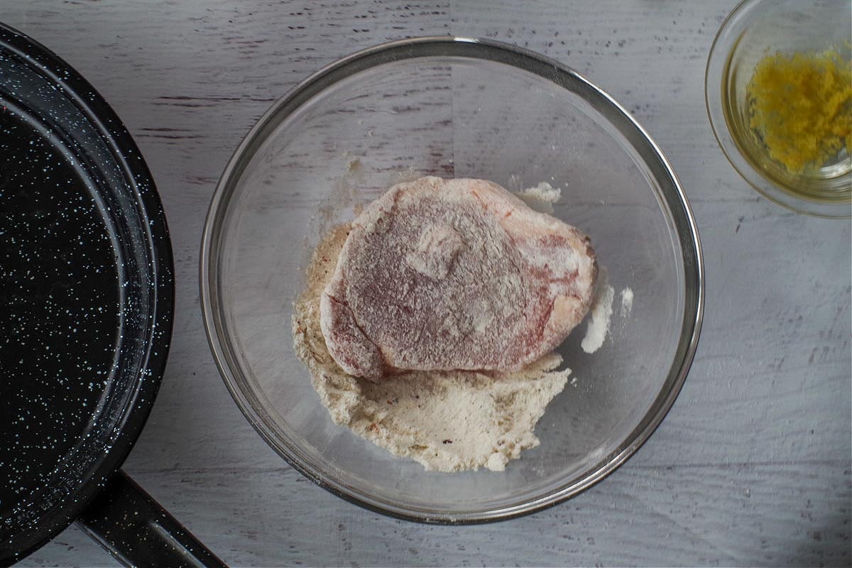 A piece of pork being coated with flour mixture in medium glass bowl.