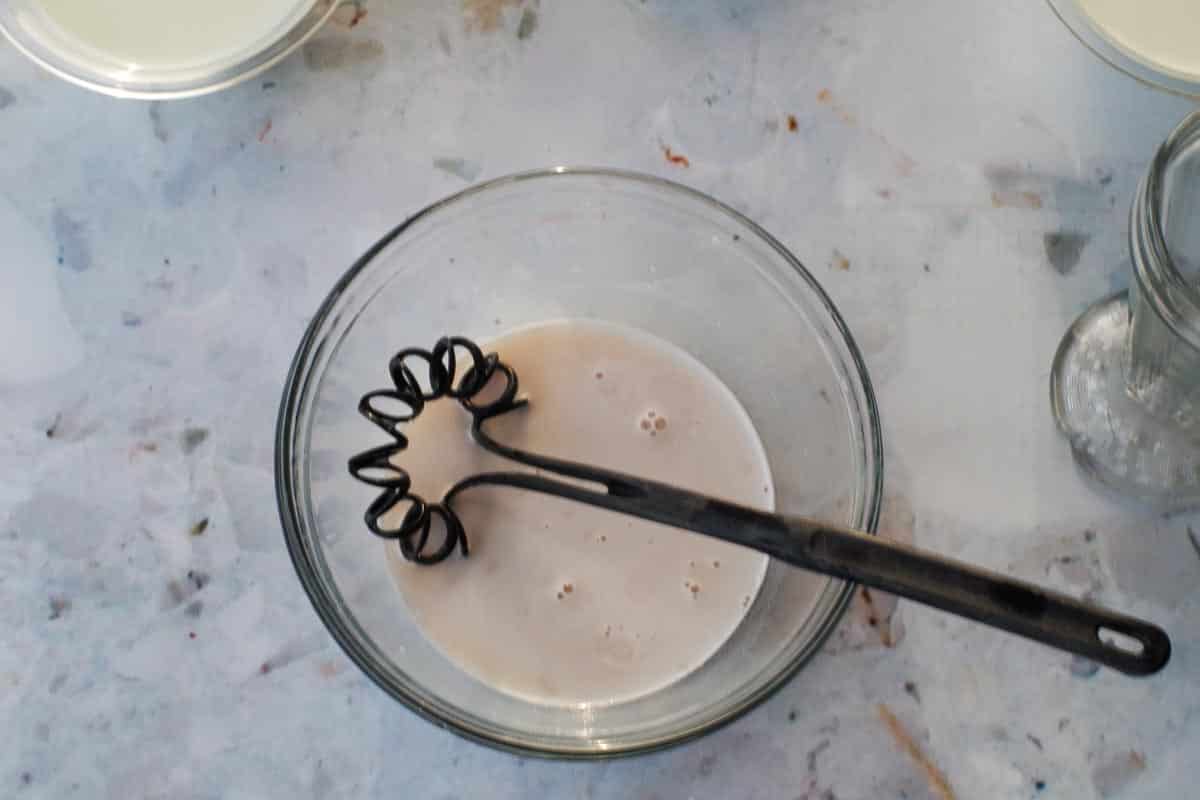 Boiling hot water and white chocolate mix in a medium glass bowl with a black whisk.