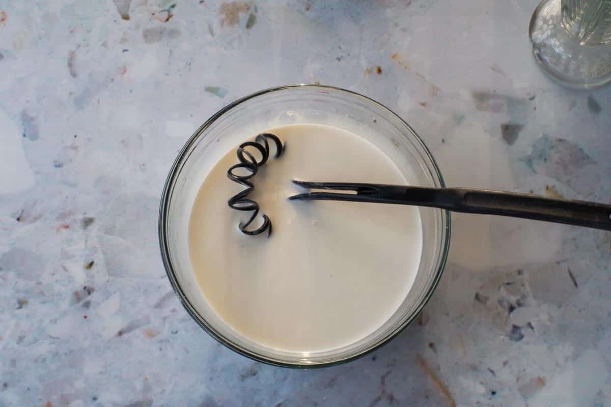 Milk, half and half cream and extracts added to white hot chocolate in a glass bowl with a whisk.