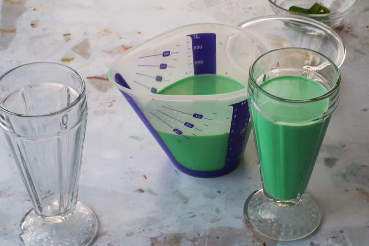 A large measuring cup ⅓ full of green grasshopper mocktail and a milkshake glass ⅔ full, beside and empty cup. 