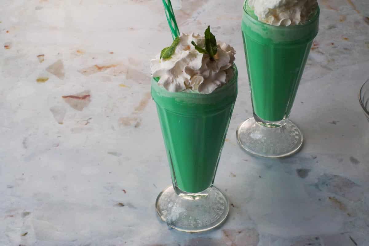 Green Grasshopper Mocktail in a Milkshake glass with whipped cream and mint leaves garnish and a green straw, with another same drink in the background.