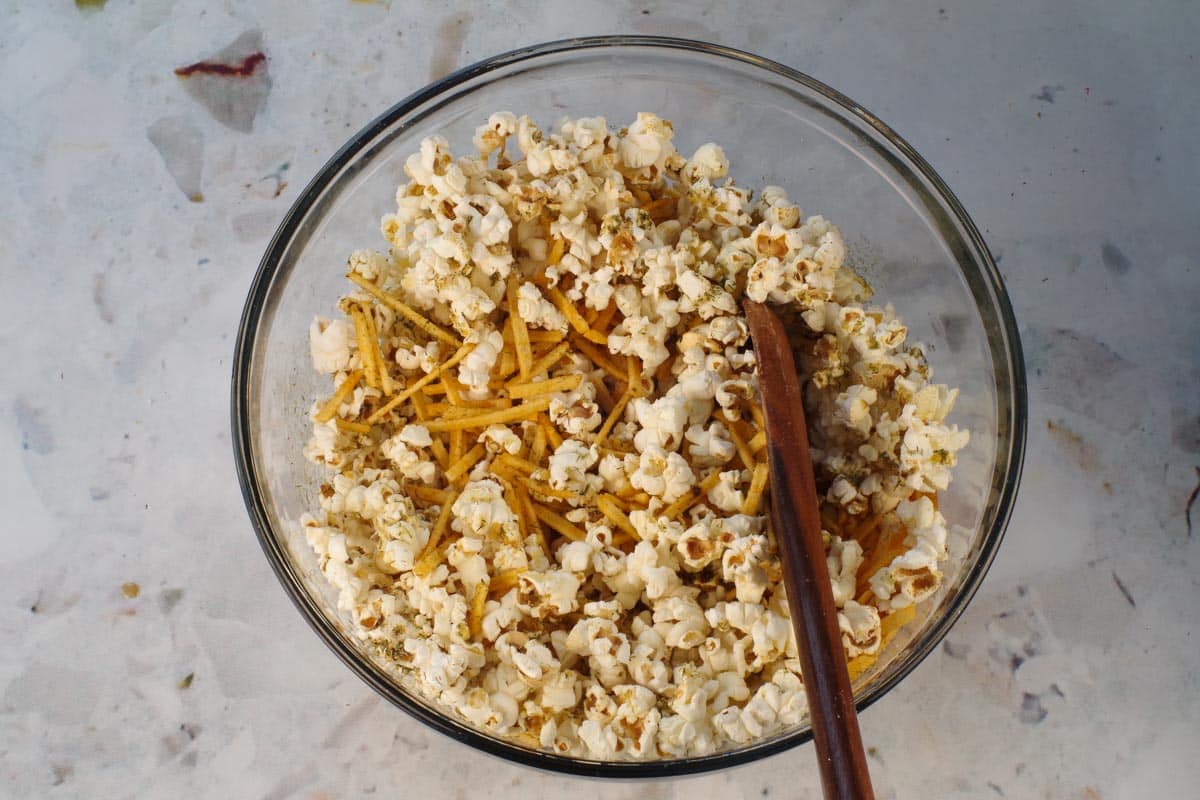 Popcorn being mixed together in a large glass bowl with a wooden spoon after remaining butter mixture and spices are added.