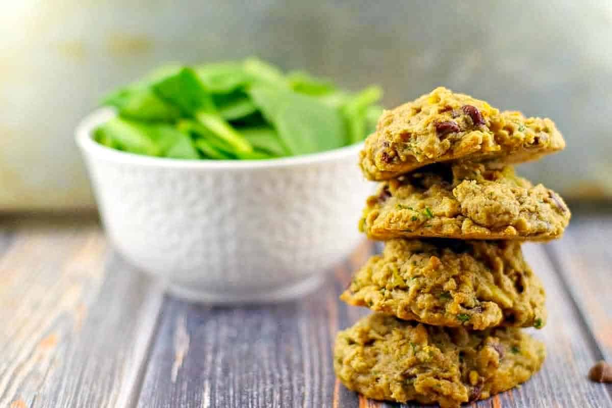 4 healthy chocolate spinach cookies stacked with a bowl of fresh spinach in the background.