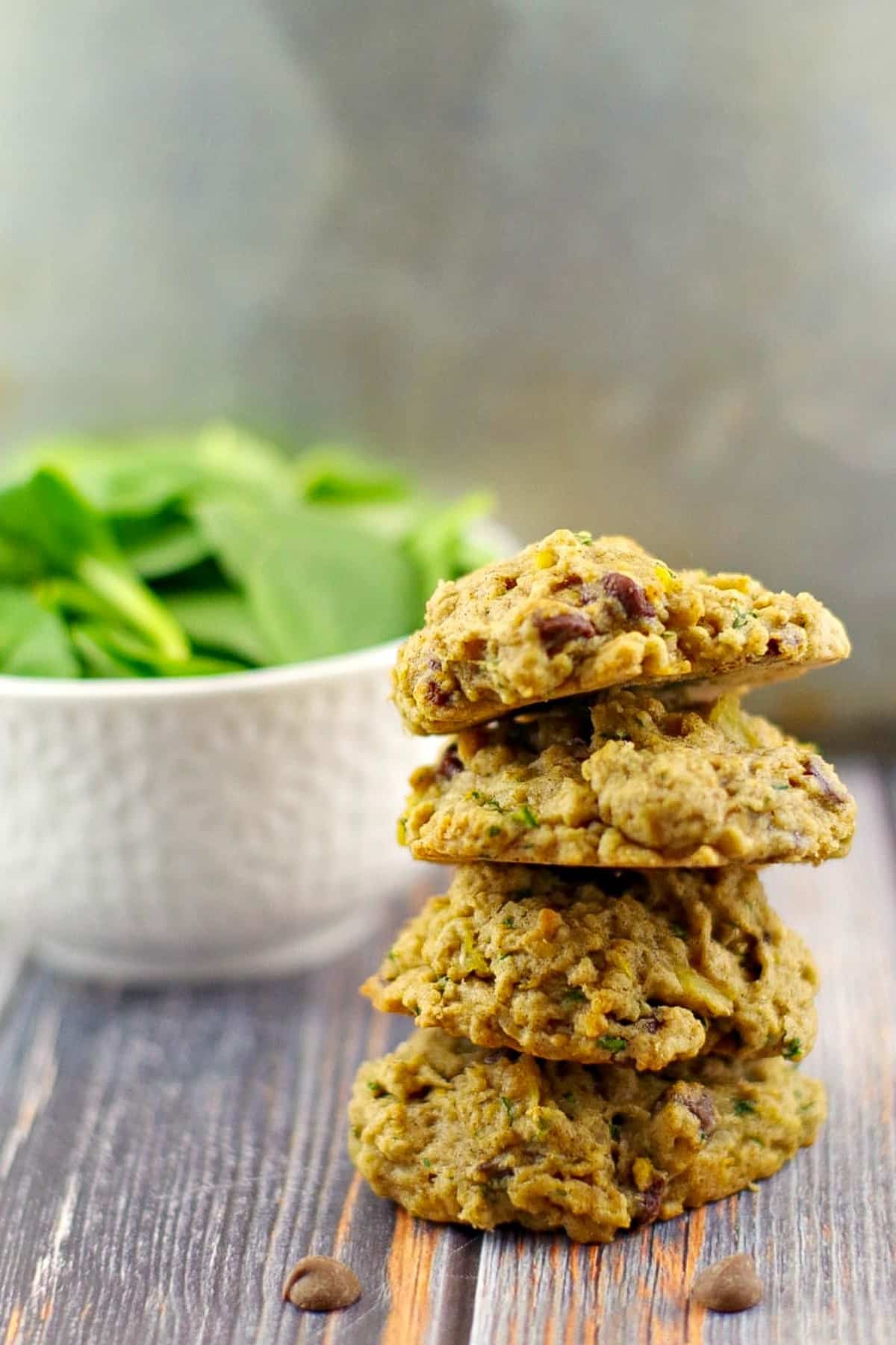 4 healthy chocolate spinach cookies stacked with a bowl of fresh spinach in the background.