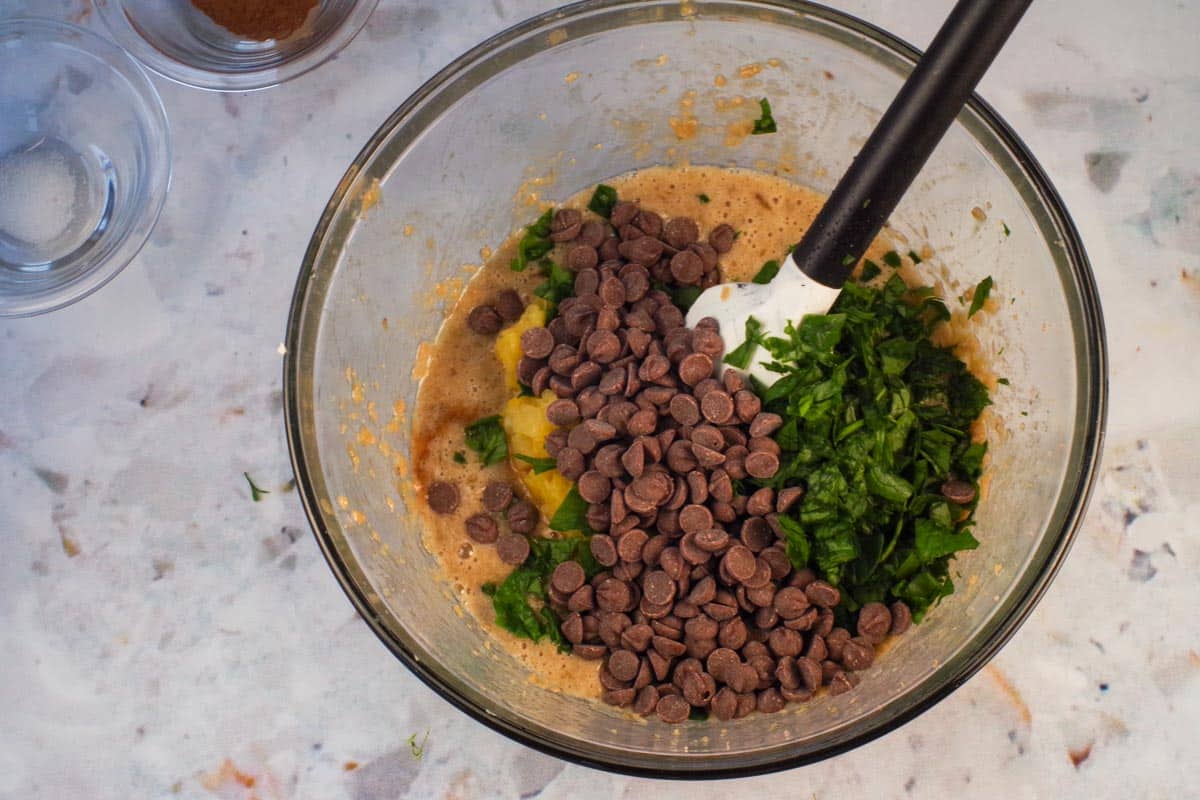 Spinach,, pineapple and chocolate chips added to mixture in large glass bowl with spatula.