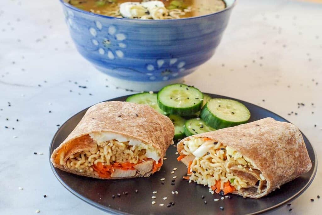 Ramen wrap on a black plate with cucumbers and ramen soup in the background.